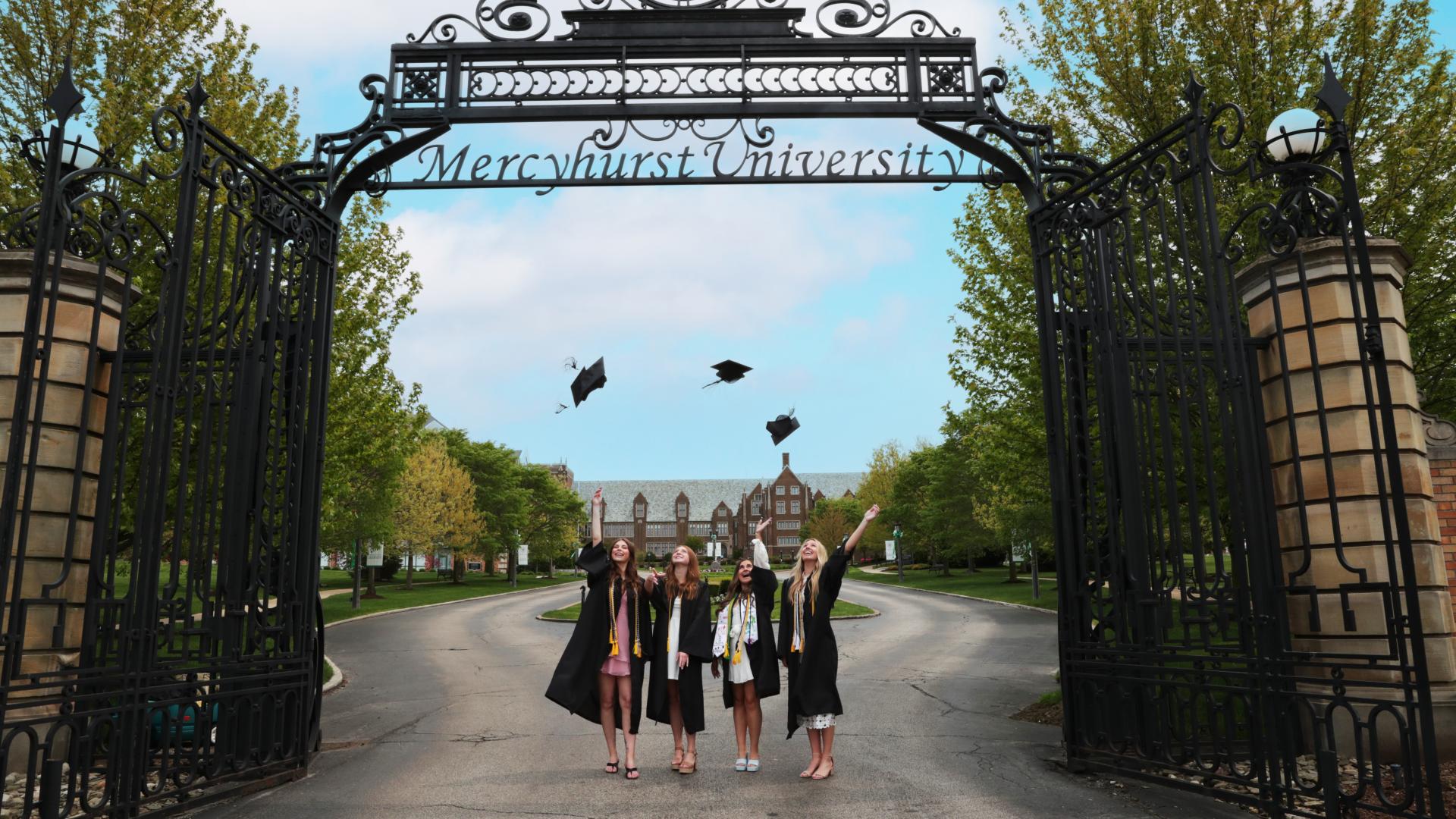 four female Ƶapps throw graduation caps under the mercyhurst gates